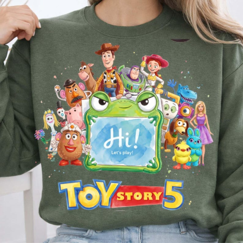May include: An olive green crewneck sweatshirt featuring a colorful graphic of various Toy Story characters surrounding a central green frog-shaped frame. The frame contains the text Hi! Let's play! Below the characters, the text TOY STORY 5 is printed.
