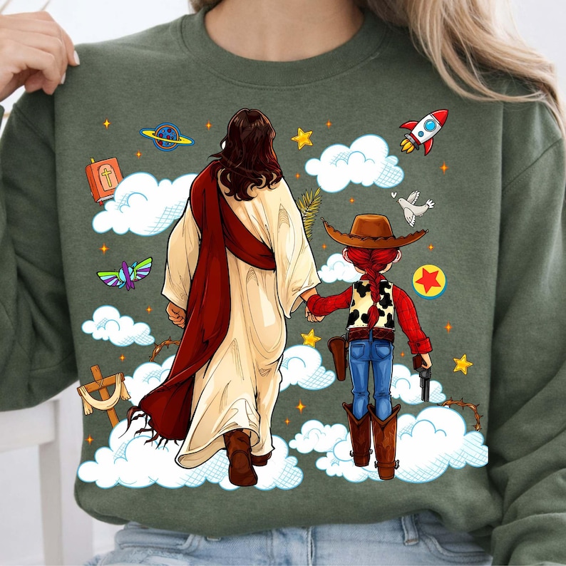 May include: Green sweatshirt featuring a graphic of Jesus holding hands with a cowgirl. The design includes clouds, stars, a rocket, and other religious and cartoon elements. The cowgirl wears a brown hat, vest, and boots.
