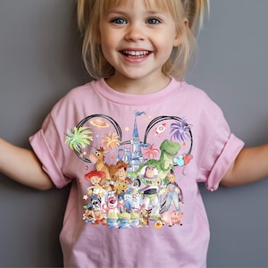 May include: Pink t-shirt featuring a colorful graphic of Disney's Toy Story characters. The design includes Woody, Buzz Lightyear, Rex, and other characters, set against a castle and palm trees within Mickey Mouse ears.