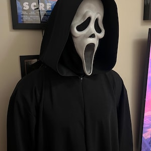 Pode incluir: Uma fantasia preta com capuz e uma máscara Scream branca. A máscara apresenta um design de rosto distorcido e gritando. A fantasia tem zíper na frente e é exibida em ambientes internos.