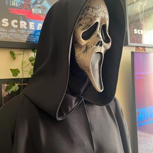 Könnte beinhalten: Eine Ghostface-Maske aus dem Film Scream mit einem verwitterten Finish, getragen mit einem schwarzen Kapuzenmantel. Die Maske hat ein blasses, schädelähnliches Aussehen mit dunklen Augenhöhlen und einem klaffenden Mund. Der Umhang ist aus glattem, dunklem Stoff.