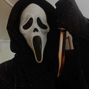 Könnte beinhalten: Eine Scream-Maske mit einem blassen Gesicht und schwarzen Augenhöhlen und Mund. Die Maske ist Teil eines schwarzen Kapuzen-Kostüms. Ein goldfarbenes Messer wird vor dem Kostüm gehalten.