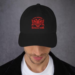 Puede incluir: Gorra de béisbol negra con un gráfico rojo. El gráfico incluye un águila bicéfala, pesas y los textos "GYM APPAREL" e "ILLYRIAN TRIBE". La gorra tiene una visera curva y una correa ajustable.