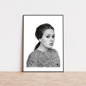 May include: Monochrome portrait print of a woman with dark hair styled in a ponytail. She wears a patterned top. The print is framed in black and displayed on a white wall, above a wooden floor.