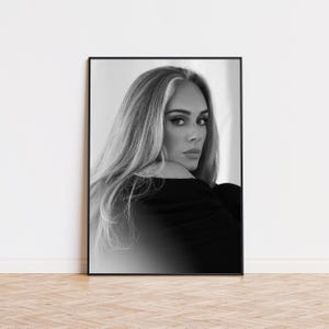 May include: Black and white framed portrait of a woman with long blonde hair. The portrait is a close-up shot, with the woman looking over her shoulder. The frame is black, and the background is a neutral color.