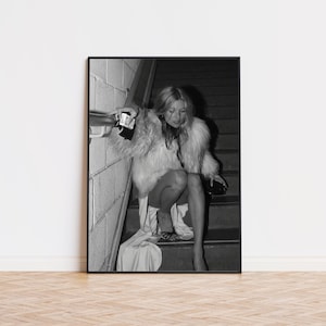 Può includere: Stampa incorniciata in bianco e nero raffigurante una persona con un cappotto soffice e chiaro, seduta su una scala. La persona tiene in mano una piccola pochette nera e una macchina fotografica. Estetica vintage.