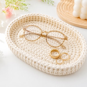 May include: An oval-shaped, cream-colored crocheted tray with a thick, textured weave. Resting inside the tray are a pair of light brown, translucent-framed eyeglasses and three gold-toned metal rings of varying thicknesses and designs.