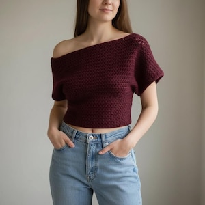 以下が含まれることがあります： バーガンディ色のオフショルダーかぎ針編みトップス、半袖。トップスはテクスチャードステッチパターンとクロップド丈です。モデルはライトブルーのジーンズを着用しています。トップスは手作りの衣服です。