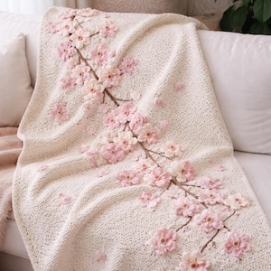 Puede incluir: Manta de punto color crema con diseño floral. La manta presenta una rama con flores y capullos rosas. La manta está sobre un sofá blanco, creando un acento decorativo y acogedor.