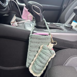 May include: A crocheted phone holder and hand sanitiser holder in shades of grey and white. The phone holder is rectangular, and the sanitiser holder is cylindrical. Both are attached to a gold-coloured keychain. The items are hanging in a car.