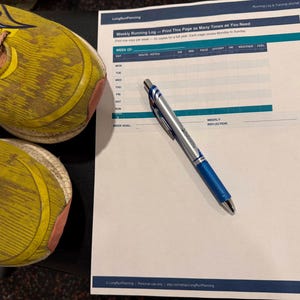 Può includere: Scarpe da corsa gialle con lacci rosa e un design blu e giallo. Un registro di corsa settimanale stampato e una penna blu sono su una superficie scura. Il registro include il testo "Weekly Running Log - Print This Page as Many Times as You Need."