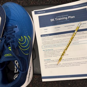 Può includere: Un paio di scarpe da corsa blu con dettagli gialli e suole bianche accanto a un piano di allenamento 5K stampato. Il piano di allenamento è su un foglio di carta bianco con intestazioni blu e viene indicato da una matita gialla.