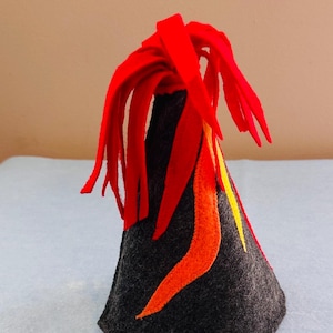May include: A cone-shaped felt volcano toy in dark gray with red felt strips attached to the top to simulate erupting lava. The sides of the volcano feature appliqued felt flames in bright orange and yellow colors against the dark gray base.