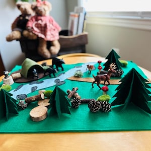 May include: A felt forest scene with a green base, featuring a river, trees, and miniature animal figurines. Includes a tent, logs, and pine cones. The scene is set on a wooden table, with teddy bears in the background.