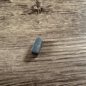May include: A small, dark gray, rectangular object with a slightly textured surface. The object is lying on a wooden surface with a natural wood grain pattern.