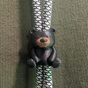 May include: A black bear-shaped cord lock slider featuring a brown snout, threaded onto two white and black patterned drawstrings. The cord lock is positioned against an olive green fabric background, serving as a functional fastener for the cords.