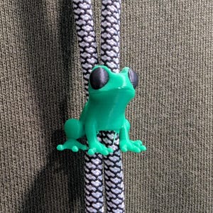 May include: A bright green frog-shaped cord lock attached to two black and white patterned drawstrings. The frog has large black eyes and is positioned against a textured olive green fabric background, serving as a functional toggle.