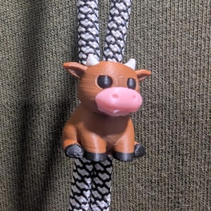 May include: A brown cow figurine with a pink snout, white horns, and black hooves attached to two black and white patterned cords. The cow has large black eyes and a rounded body, serving as a decorative cord lock or slide for a hooded garment.