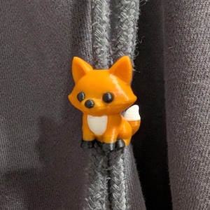 May include: A small, orange fox-shaped cord lock attached to two grey braided drawstrings on a dark grey fabric garment. The fox figure features a white chest patch, a white-tipped tail, black eyes, and black paws, serving as a functional toggle.