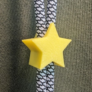 May include: A bright yellow star-shaped cord lock made of plastic, featuring a five-pointed design. The cord lock is threaded onto two parallel black and white braided drawstrings, which are positioned against a textured olive green fabric background.