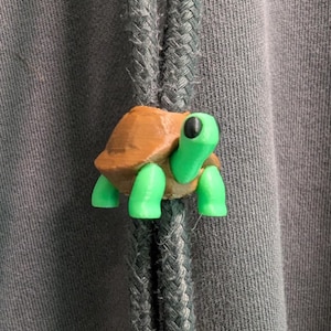 May include: A small, three-dimensional turtle-shaped drawstring toggle attached to two thick, grey braided cords. The turtle features a brown shell and a bright green head, neck, and four legs, with a solid black eye visible on the side of its head.