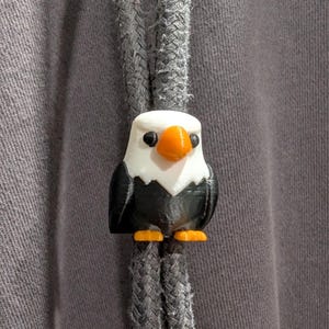 May include: A gray hoodie drawstring cord featuring a 3D printed bald eagle bead. The eagle has a white head, black body, orange beak, and orange feet. The bead is positioned centrally on the two gray braided cords of the garment.