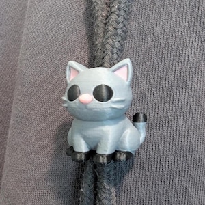 May include: A gray cat-shaped drawstring toggle attached to two dark gray braided cords. The cat figure has large black eyes, a small pink nose, pink inner ears, and black paws and tail tip, secured onto the fabric of a dark gray hooded garment.