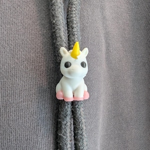 May include: A small white unicorn figurine with a yellow horn and pink hooves attached to a dark gray drawstring cord. The unicorn has large black eyes and is positioned vertically between two thick, textured gray fabric hoodie strings.