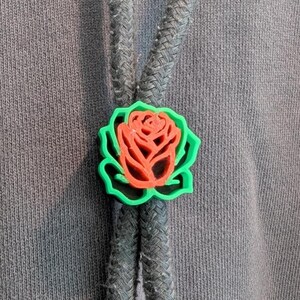 May include: A bolo tie featuring a red rose design with a green outline. The rose is set against a gray braided cord, with a gray fabric background. The bolo tie is a unique accessory.
