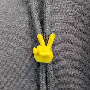 May include: A bright yellow peace sign hand gesture charm on a gray drawstring. The charm is a three-dimensional design, with the fingers clearly defined. The background is a textured gray fabric.