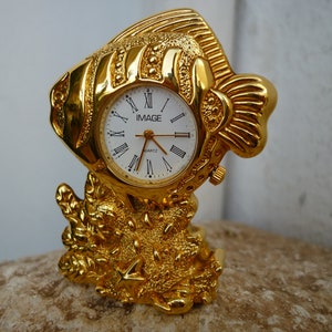 Könnte beinhalten: Goldfarbener Tischuhr in Form eines Fisches mit weißem Zifferblatt und römischen Ziffern. Die Uhr steht auf einem goldfarbenen Sockel, der an Korallen erinnert.