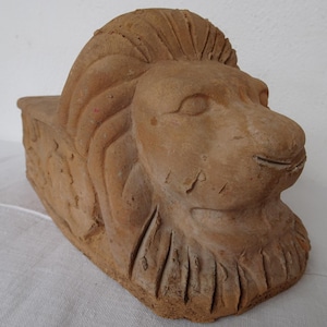 Puede incluir: Escultura de león de terracota con una melena y rasgos faciales detallados. La cabeza del león está esculpida con una expresión serena, y la pieza tiene una apariencia envejecida. La escultura es probablemente un objeto decorativo.
