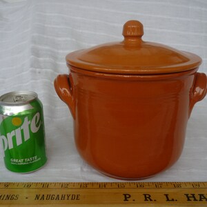 May include: A glazed, orange-brown ceramic pot with a matching lid and two side handles. A green and silver Sprite can is next to the pot for size comparison. The pot is likely for cooking or storage.