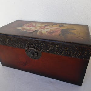 Puede incluir: Caja rectangular de madera con tapa con bisagras. La tapa está pintada con un diseño floral en tonos rosa, amarillo y marrón. La caja tiene un borde decorativo marrón oscuro y un cierre de metal.