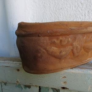 Könnte beinhalten: Ein Terrakotta-Pflanztopf mit einem dekorativen Design aus stilisierten Blättern und Blumen. Der Topf ist braun und hat eine leicht verwitterte Optik.