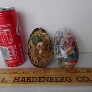 Puede incluir: Una lata roja de Coca-Cola junto a dos latas decorativas de huevos de Pascua. Una lata contiene un pequeño conejito de peluche en un nido de paja. La otra lata tiene una escena pintada de un conejo con una cesta. Una regla de madera está en primer plano.