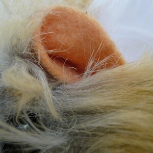 May include: Close-up of a plush toy's ear, featuring a soft, peach-colored fabric. The ear is surrounded by fluffy, light brown and gray fur.