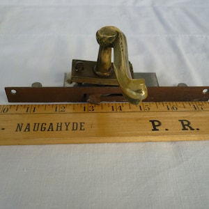 Könnte beinhalten: Ein Türverschluss aus Messing mit einem dekorativen Griff. Der Verschluss ist auf einer Metallplatte montiert und an einem Holzliniensystem befestigt, das in Zoll gekennzeichnet ist. Das Lineal ist mit "INGS - NAUGAHYDE" und "P.R.L.I." beschriftet.