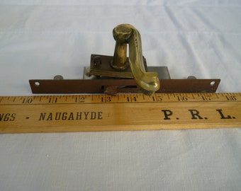 May include: A brass door latch with a decorative handle. The latch is mounted on a metal plate and is attached to a wooden ruler marked with inches. The ruler is labeled "INGS - NAUGAHYDE" and "P.R.L.I."