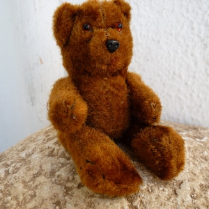 May include: A vintage brown teddy bear with red eyes and a black nose. The plush toy is sitting on a textured, light brown surface. The bear has a classic design with visible stitching and a soft, furry texture.