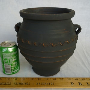 May include: A large, dark gray ceramic pot with two handles and a decorative wavy pattern. The pot has a wide opening and a rounded body. A green soda can is next to the pot for size comparison.
