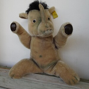 Op de afbeelding: Een vintage pluche neushoorn speelgoed in bruin- en beigetinten. De neushoorn heeft een zwarte manen, een prominente hoorn en donkerbruine accenten op zijn voeten en oren. Een geel label is aan zijn oor bevestigd.