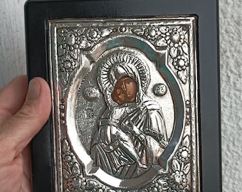 Puede incluir: Un icono religioso plateado que representa a la Virgen María sosteniendo al niño Jesús. El icono está enmarcado en un marco de madera negro.