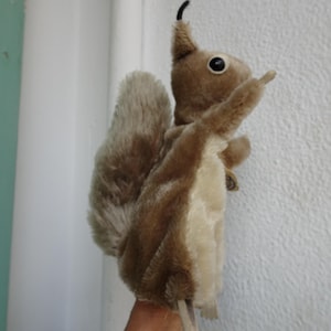 May include: A plush squirrel hand puppet featuring soft brown fur on the back and a lighter cream-colored belly. The puppet has a large, bushy tail, dark eyes, and a small black tuft on the top of its head, designed to be worn on the hand.