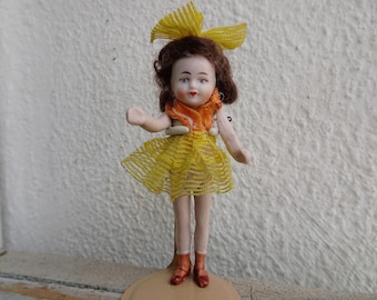May include: A porcelain doll with a yellow dress and a yellow bow in her hair. She is wearing brown shoes and is standing on a white base.