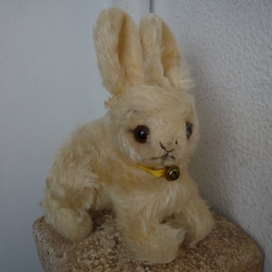 May include: A vintage cream-colored stuffed rabbit with long ears and dark eyes. It has a yellow collar with a small bell. The plush toy is sitting on a stone surface.