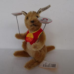 May include: A plush kangaroo toy with a red vest and yellow trim, holding two small plates on wires. The kangaroo has a tag attached to its ear and a round tag on its vest. The toy is brown and tan.