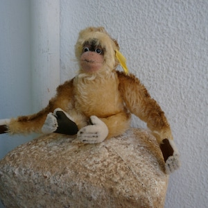 May include: A vintage stuffed monkey with light brown fur and a cream-coloured face, hands, and feet. The monkey has dark eyes, a pink nose, and is seated on a light-coloured stone. A yellow ribbon is attached to its head.