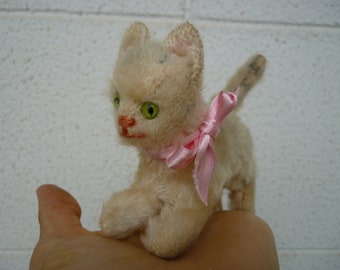 May include: A white plush toy cat with green eyes and a pink bow. The cat is sitting on a hand.