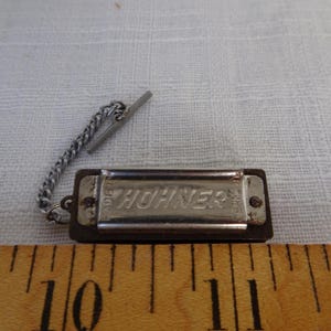 May include: A small Hohner harmonica with a silver metal cover and the brand name "Hohner" embossed on it. It has a dark brown body and a silver chain attached to a silver bar. A wooden ruler is in the foreground.